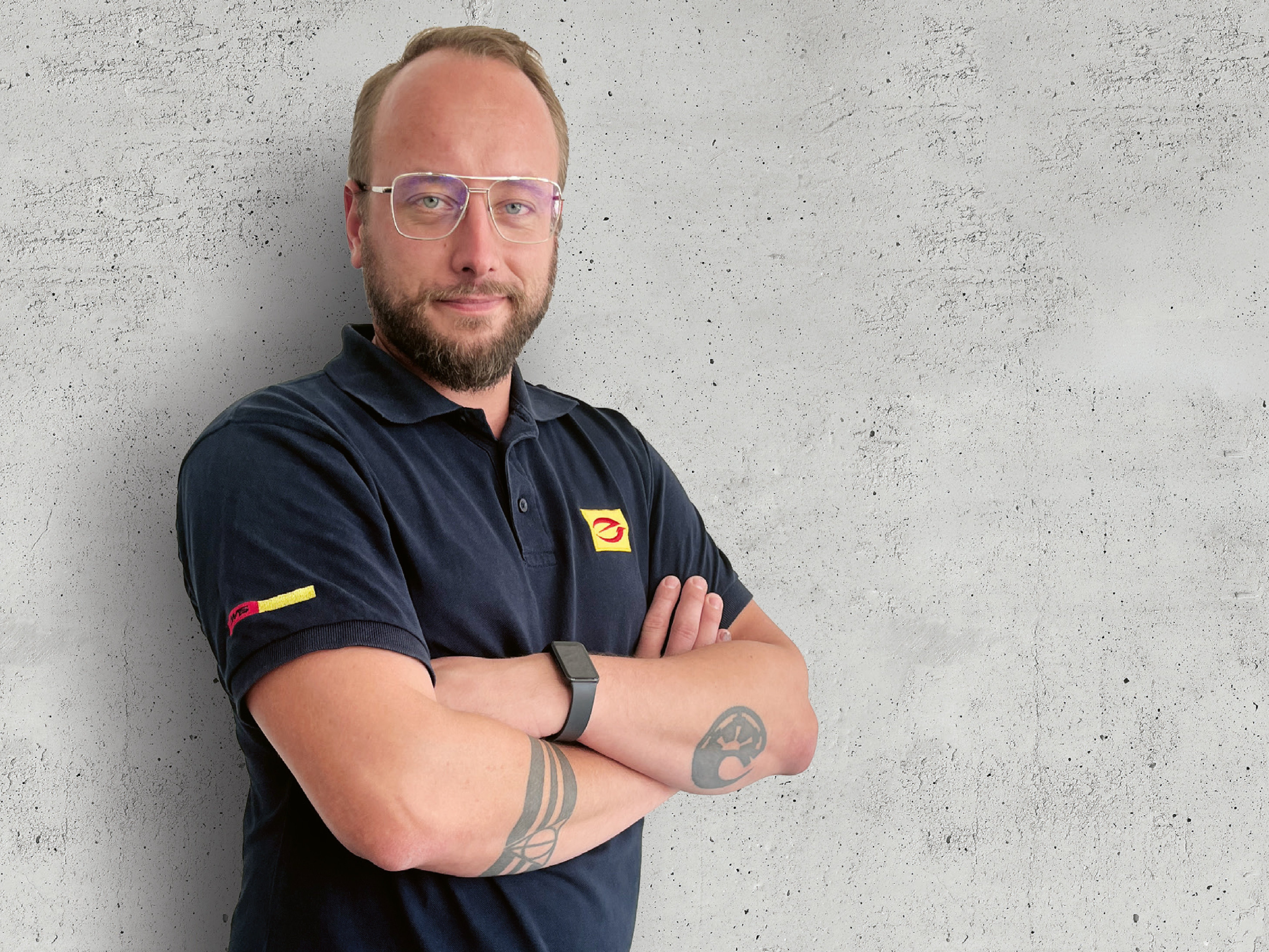 Technischer Berater steht mit verschränkten Armen vor grauer Betonwand. Er trägt ein schwarzes Poloshirt mit gelb-rotem Logoaufdruck.