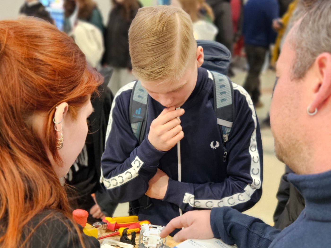 Junger Besucher mit Rucksack und dunkelblauer Jacke betrachtet den Mitmachsockel und lässt sich zeigen, wie man eine Steckdose richtig anschließt.