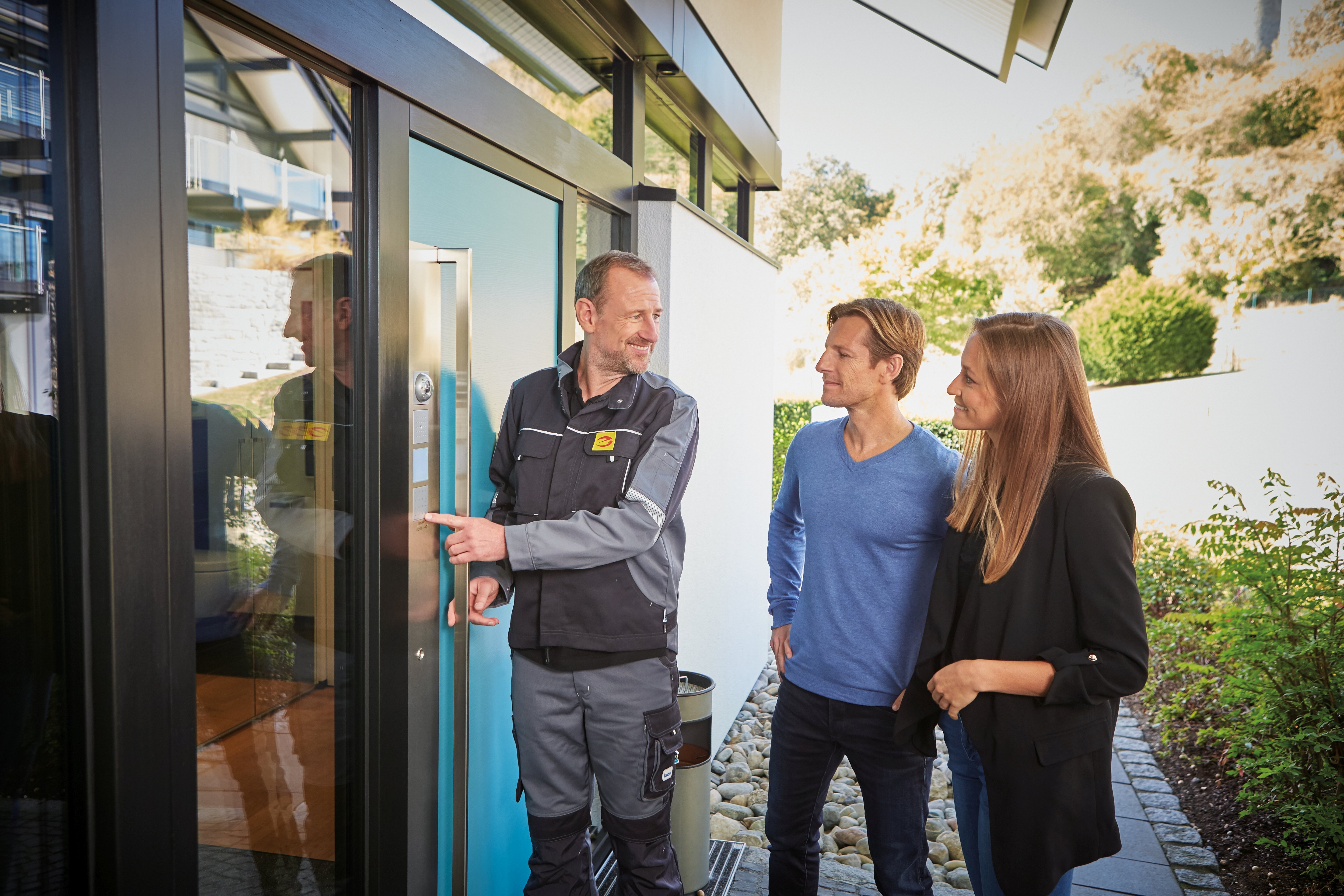 Ein Mann in Arbeitskleidung mit "e-Marke"-Aufdruck zeigt seinen Kunden vor einem modernen Glaseingang die Funktionalität des eingebauten Sicherheitssystems.
