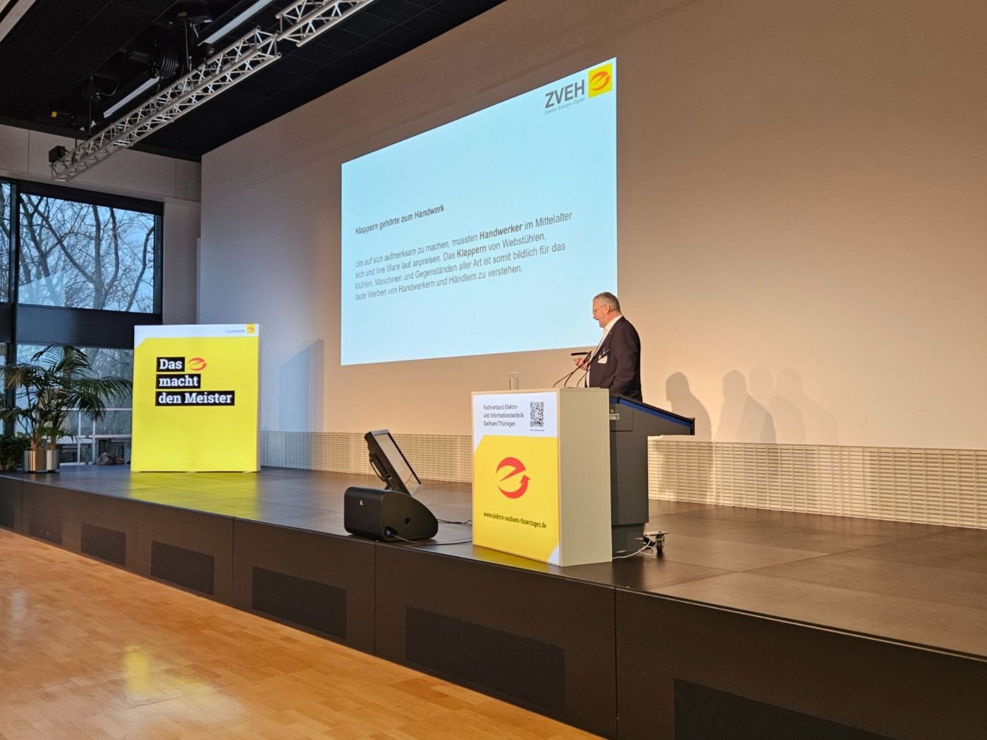 Blick in den Veranstaltungsraum der Niederspannungsfachtagung in Dresden. Der Referent steht auf der Tribune und hält seinen Vortrag. Hinter ihm ist eine Leinwand, auf der die Präsentation dargestellt wird.