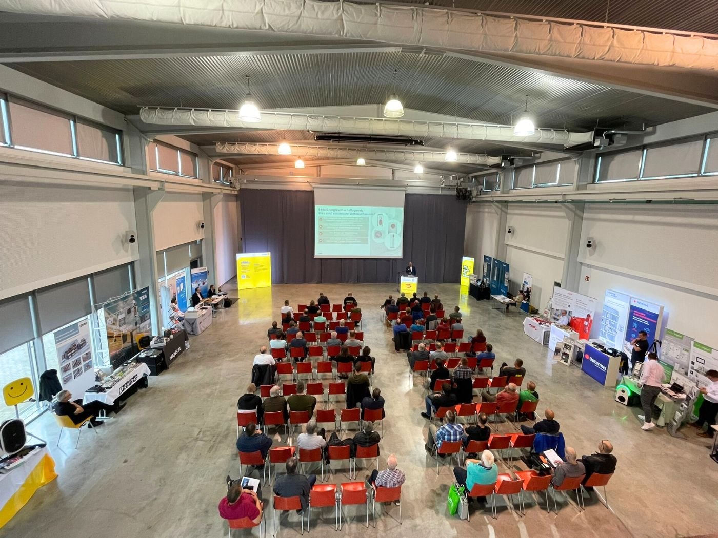 Bild mit Blick in den Veranstaltungsraum der Fachschulungstage 2024 in Nohra/Weimar. Auf der Tribüne steht ein Referent, hinter diesem ist eine Folie einer Powerpoint-Präsentation auf der Leinwand abgebildet.