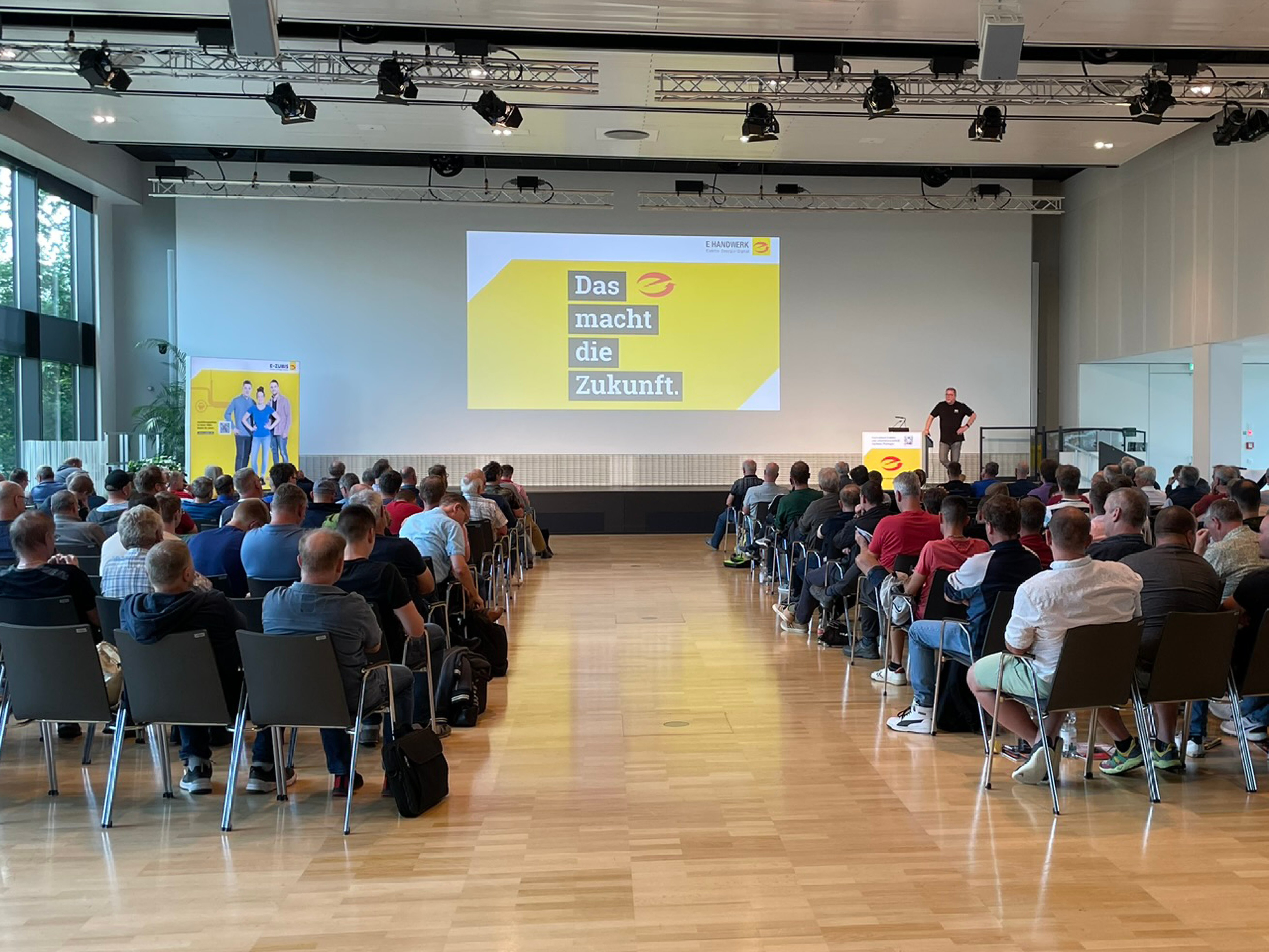 Großer Saal mit Publikum auf Stühlen, vorne befindet sich eine Bühne mit Redner und großer Leinwand, auf der "Das e macht die Zukunft." steht.
