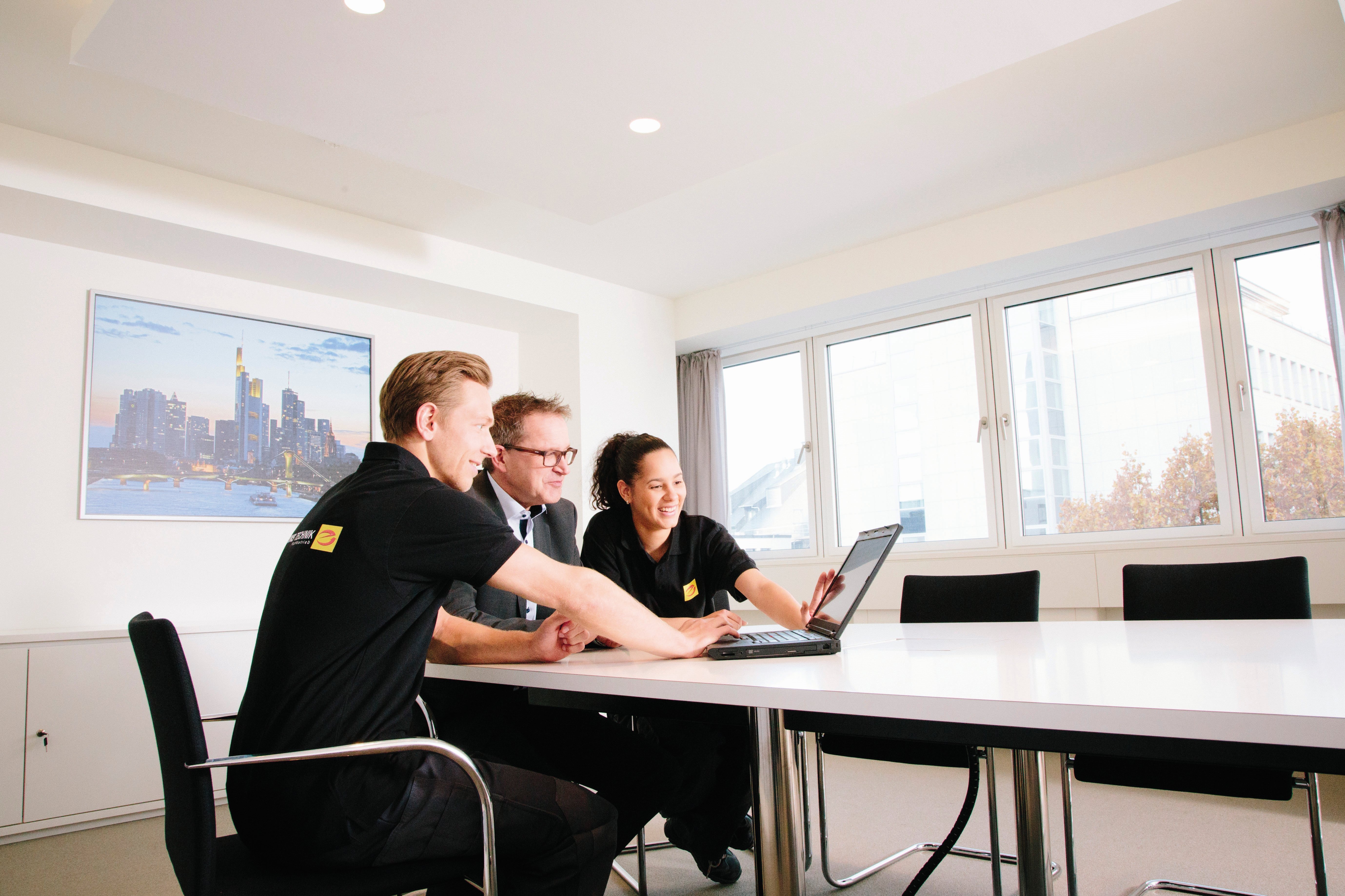 Drei Personen sitzen an einem weißen Konferenztisch und schauen auf einen Laptop, im Hintergrund befindet sich ein Bild einer Stadtansicht an einer Wand.