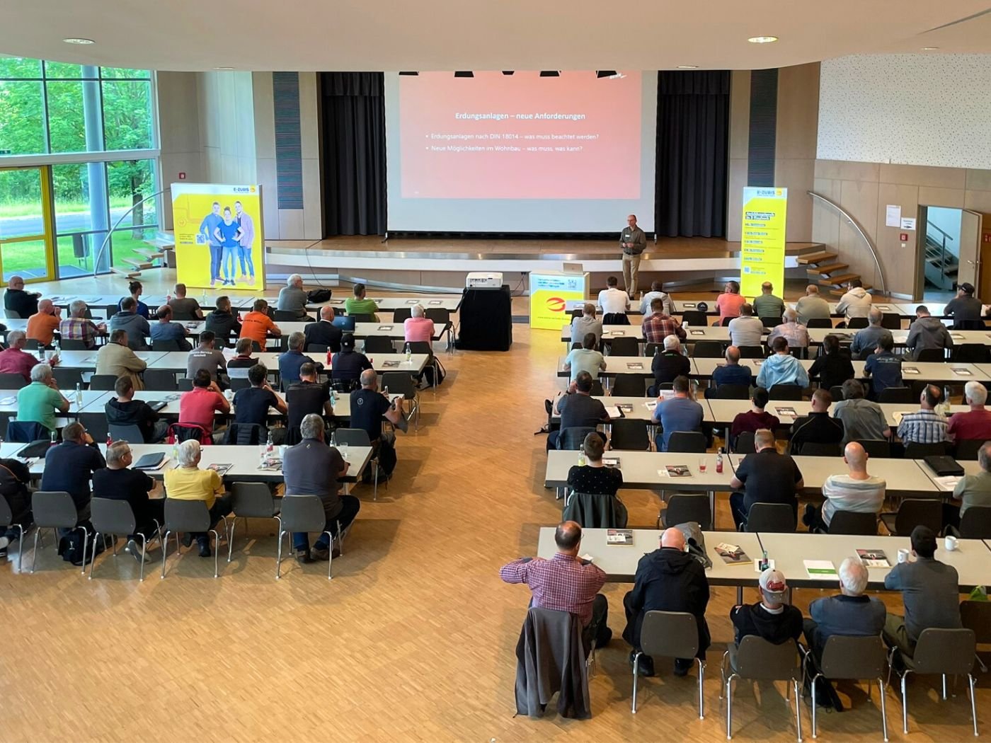Bild mit seitlichen Blick in den Veranstaltungsraum der Fachschulungstage 2024 in Glauchau. Auf der Tribüne ist eine Folie einer Powerpoint-Präsentation auf der Leinwand abgebildet.