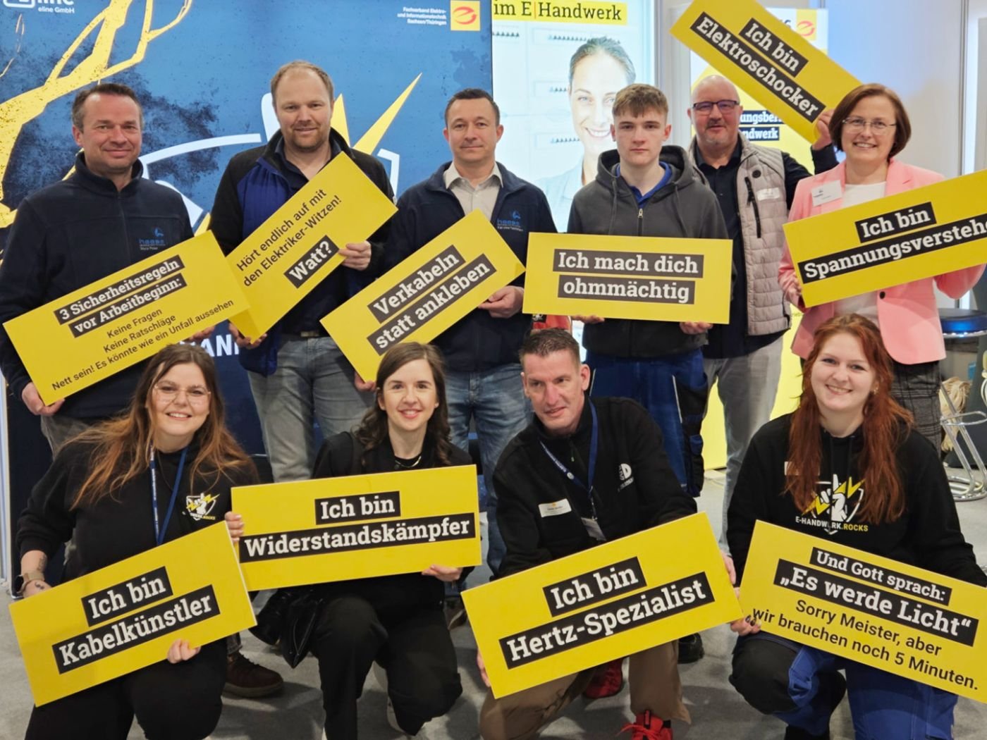 Gruppenfoto von Messeteam 2026. Alle halten gelbe Schildern mit Sprüchen zu Elektrotechnik und Handwerk in die Kamera.