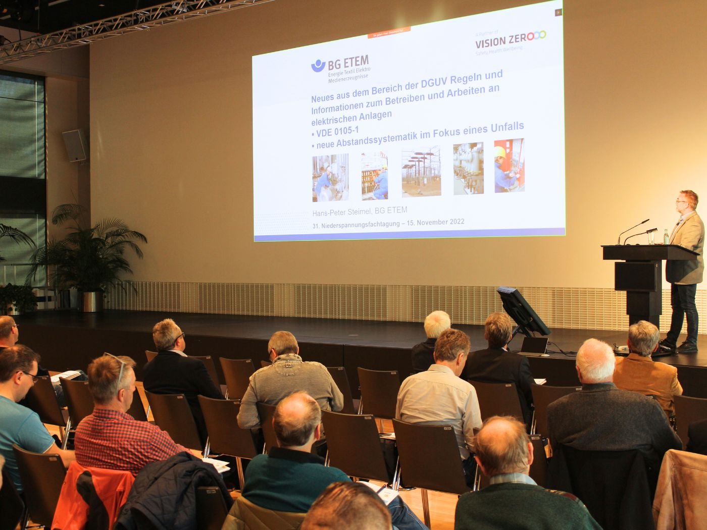 Blick in den Veranstaltungsraum der Niederspannungsfachtagung in Dresden. Der Referent steht auf der Tribune und hält seinen Vortrag. Hinter ihm ist eine Leinwand, auf der die Präsentation dargestellt wird.